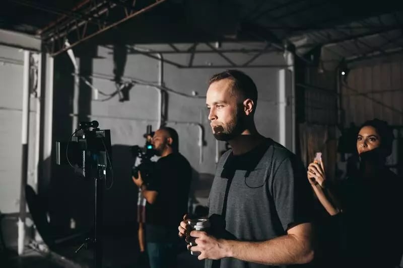 A camera man wearing a black shirt and holding a cup of coffee looks at a camera setup on a video production set
