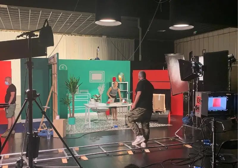 On set at a Skeleton Video Production shoot for Europa. The backdrop is green and red. In the foreground we see camera equipment, in the background we see a producer talking to the actor.