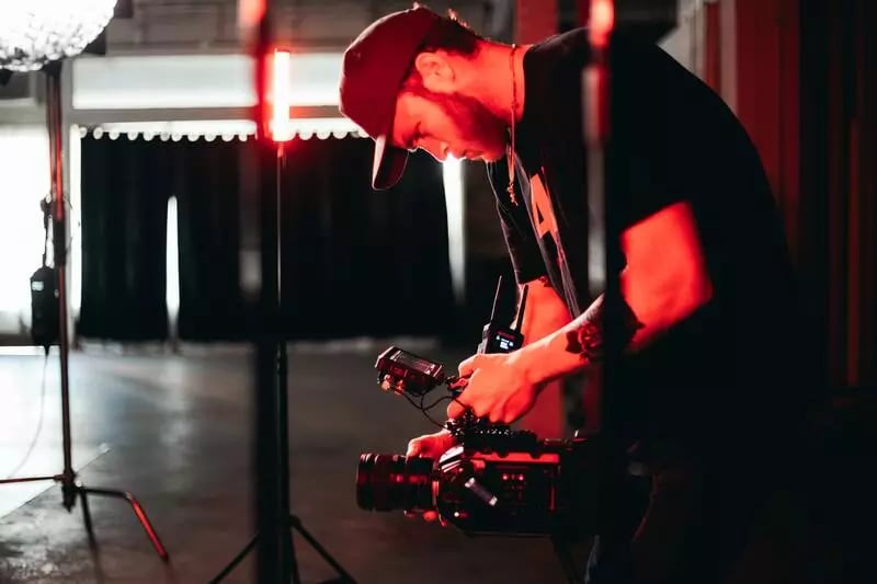 A camera man wears a cap and holds a camera while bending down. There is a red glowing light that envelopes him.