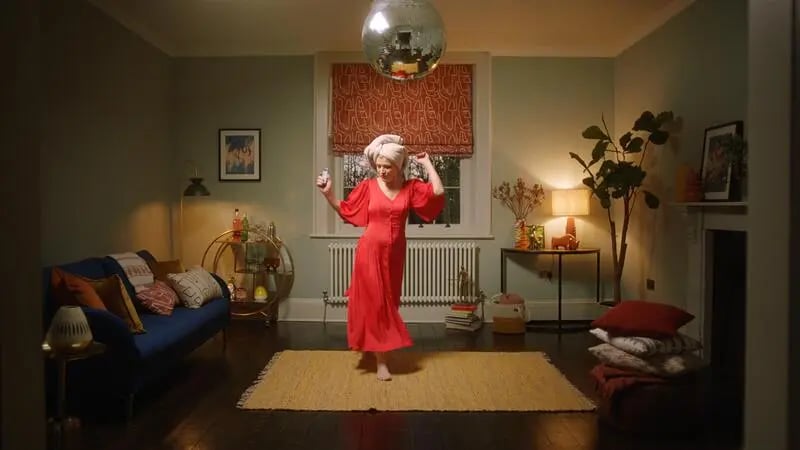 A camera scene depicting an actor in a living room set wearing a red dress and hair towel having a dance between scene takes. 