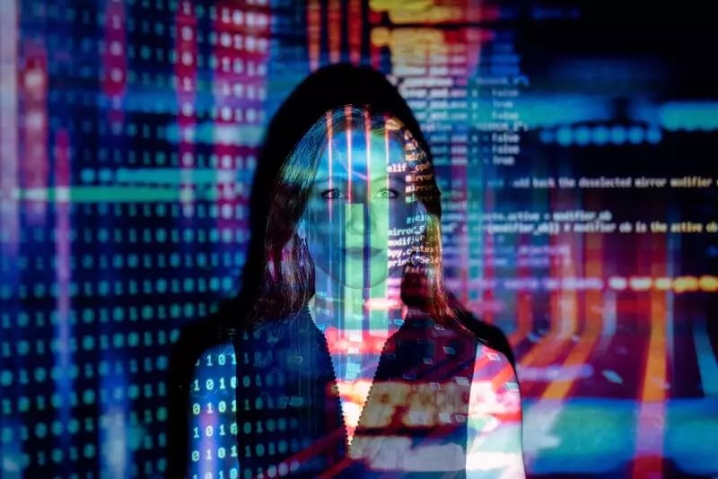A woman with blonde hair stands in font of a projector. She is bathed in colourful code.