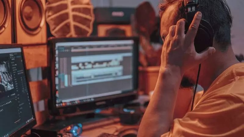 A video editor with headphones on sits at two computer screens editing a video.