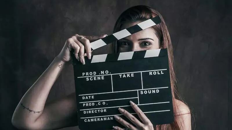 A woman with brown air hides her face behind a clapper on a video production company set against a grey backdrop